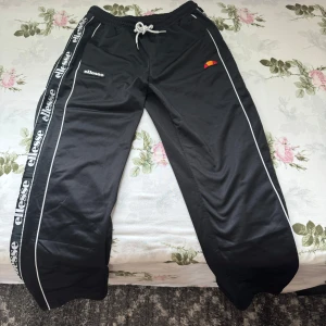 Svarta byxor från Ellesse - Svarta byxor från Ellesse med vita detaljer och tryckta loggor längs sidorna. Byxorna har knappar längs benen men jag har sytt igen längs vägen så man inte ser benen igenom (sömmarna kan sprätas bort). Snörning i midjan och är gjorda i ett glansigt syntetmaterial. Perfekta för en sportig och avslappnad look.
