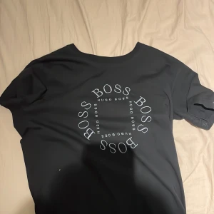 Svart t-shirt från Hugo Boss - Svart t-shirt från Hugo Boss med rund hals och vit BOSS-logga tryckt i cirkel på bröstet. Klassisk passform och korta ärmar, perfekt för en clean och stilren look. Materialet känns mjukt och skönt mot huden.