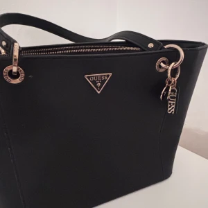 Svart handväska från Guess - Snygg svart handväska från Guess med guldfärgade detaljer och dragkedja. Väskan har klassisk form, två handtag och en Guess-logga framtill. Insidan är ljusbeige och har märkets tryck. Perfekt för dig som vill ha en stilren och trendig accessoar.