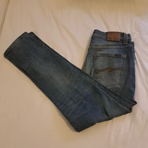 Ljusblå jeans från Nudie Jeans - Säljer ett par ljusblå jeans från Nudie Jeans med klassisk femficksdesign och snygga kontrastsömmar. Jeansen har raka ben och subtila slitningar framtill. Bakfickorna har Nudie Jeans signaturbrodyr i orange. Perfekta för en avslappnad stil. Köpt för 500