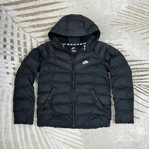 Svart pufferjacka från Nike - Puffig och varm jacka från Nike, perfekt för höst och vinter. Unisex-modell i storlek 158–170 (passar både ungdom och mindre vuxen beroende på passform). Tillverkad i 100% polyester, vilket gör den både lätt och isolerande. Lite sliten med några fläckar, men annars i fint skick och fullt användbar. En bra jacka för kyligare dagar!