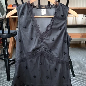 Svart linnetopp med spets Isabell Kristensen - Supersnygg svart linnetopp från Isabell Kristensen med v-ringning och spetsdetaljer längs halsringningen och midjan. Toppen är dekorerad med små svarta pärlor och broderade stjärnor, vilket ger en lyxig känsla. Perfekt för dig som gillar unika detaljer och elegant stil.