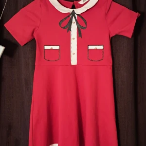 Röd kortklänning med rosett från H&M (barn) - Supersöt röd kortklänning från H&M med vit krage, svarta rosettdetaljer och fejkfickor framtill. Klänningen har korta ärmar, dekorativa knappar och en liten öppning med knapp i nacken. Den är för barn i åldern 6-8 år, och passar perfekt till julfirande :)