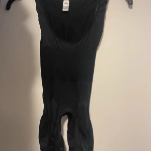 Svart sömlös jumpsuit L-XL - Svart, tight och sömlös jumpsuit med djup urringning och ribbad midja. Stretchigt material som sitter skönt och formar kroppen. Perfekt för dig som vill ha en clean och enkel look med maximal komfort.