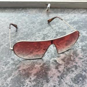 Röda shield-solglasögon från Vasuma - Snygga shield-formade solglasögon från Vasuma med röda tonade glas och tunn metallbåge. Skalmarna har ett unikt mönster i brunt och guld, vilket ger en cool retrovibe. Perfekta för dig som vill sticka ut med en statement-accessoar.