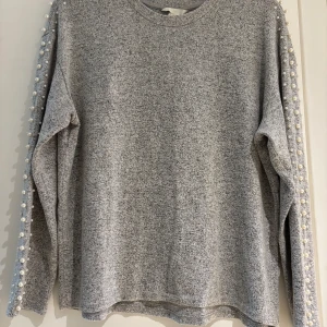 Grå tröja med pärldetaljer från H&M - Mysig grå tröja från H&M med pärlor längs axlar och ärmar. Långärmad och i mjukt material, perfekt för lager-på-lager. Enkel men ändå extra tack vare pärldetaljerna som ger en cool touch till din outfit.