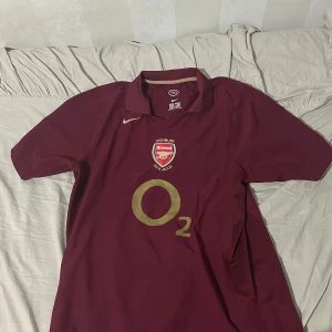 Arsenal Highbury Henry 14 Nike tröja - Säljer en ikonisk Arsenal Highbury 2005-2006 matchtröja i vinröd med guldiga detaljer. Tröjan har korta ärmar, klassisk krage och tryck med Henry 14 på ryggen. Broderat Arsenal-märke och Nike-logga framtill. Perfekt för dig som älskar fotbollshistoria. Är i Storlek XL men passar L också. Prisk kan diskuteras!