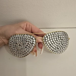 Glittriga aviator-solglasögon med stenar - Unika aviator-solglasögon med guldiga bågar och stora linser täckta av glittrande silvriga stenar. Perfekta för dig som vill sticka ut och glänsa på nästa fest eller event. Riktigt statement piece för en cool look.
