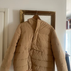 Beige anorak från Studio Total - Säljer en beige anorak/pufferjacja från Studio Total i storlek S. Jackan har huva, dragkedja och knappar framtill samt en stor ficka på magen. Fodret är orange och materialet är en mix av polyester och bomull. Perfekt för kyliga dagar och riktigt snygg till streetwear-till skidresan!