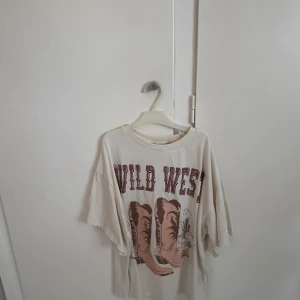 Vit oversized t-shirt Wild West - Vit oversized t-shirt med tryck framtill där det står 'WILD WEST' och motiv av cowboy boots i brunt och beige. T-shirten har korta ärmar och rund halsringning. Perfekt för dig som gillar western vibes och avslappnad stil. Stl 134-140 men passar större storlekar då den är oversized.
