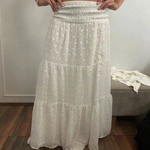 Vit långkjol från Gina Tricot - Supersöt vit långkjol från Gina Tricot med smock i midjan och ett diskret geometriskt mönster. Kjolen har volangdetaljer och är tillverkad i ett lätt och luftigt tyg, perfekt för varma dagar. Sitter bekvämt och ger en bohemisk vibe.