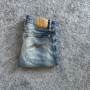 Nudie Jeans blå jeans, storlek 25/28 - Snygga blå jeans från Nudie Jeans med klassisk femficksdesign och orangea sömmar. Jeansen har en ljus tvätt och subtila slitningar för en cool, avslappnad look. Tillverkade i bomull och passar perfekt till vardagsstilen.