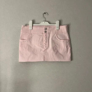 Ljusrosa jeanskjol med pärldetaljer - Supersöt ljusrosa kort jeanskjol med pärlor och silvriga nitar vid fickorna framtill. Kjolen har två knappar i midjan, bälteshällor och fickor både fram och bak. Perfekt för dig som vill ha en feminin och trendig look.