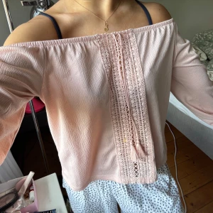 Ljusrosa offshoulder blus med spets - Superfin ljusrosa blus med offshoulder-modell och långa ärmar. Framtill har den en bred spetsdetalj med tofsar som ger en boho-vibe. Tunt, luftigt tyg som känns perfekt för varmare dagar. Snygg att styla med jeans eller kjol för en chill look.