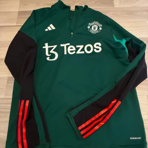 Manchester United träningströja Adidas L - Grön långärmad Manchester United träningströja från Adidas med svarta ärmar och röda ränder. Halv dragkedja framtill, klubbmärke och Adidas-logga på bröstet samt Tezos sponsortryck. Tillverkad i funktionsmaterial Aeroready.