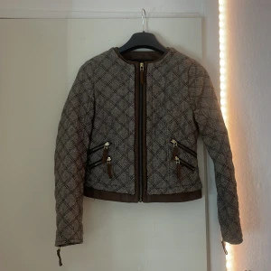 Rutig quiltad jacka från Zara Basic - Snygg rutig jacka från Zara Basic i beige och brunt med quiltad känsla. Jackan har flera dragkedjor i guld, bruna detaljer och lång ärm. Perfekt för dig som gillar en stilren men edgy look. Passar till både jeans och kjol.