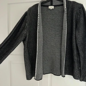 Svartvit stickad kofta från Milano - Säljer en svartvit stickad kofta från Milano i storlek M. Koftan har ett tätt, småmönstrat stickat mönster och öppen front med vit kant. Långärmad och perfekt att slänga på sig över en t-shirt eller topp för en chill look.