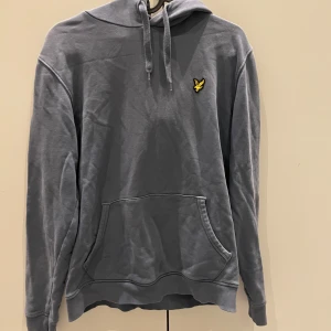 Grå hoodie från Lyle & Scott - Snygg grå hoodie från Lyle & Scott med klassisk gul logga på bröstet. Tröjan har huva med snörning och en stor magficka framtill. Perfekt för dig som gillar en enkel och stilren look. Tillverkad i mjuk bomull för skön känsla. Har använts ungefär 4 gånger och är i nytt skick, ny köpt kostar ca 1000kr