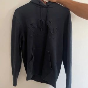 Svart hoodie från Hugo Boss - Säljer en mörkblå hoodie från Hugo Boss med stor BOSS-logga broderad på bröstet. Tröjan har huva med snörning och en stor magficka. 