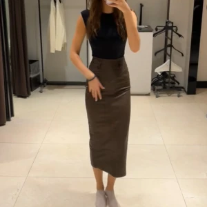 Brun lång kjol i skinnlook - Säljer en snygg brun långkjol i skinnimitation med hög midja och figurnära passform. Kjolen har bälteshällor och en diskret slits bak för extra rörelsefrihet. Perfekt för dig som gillar stilrena och trendiga plagg med edge. Från adoore 