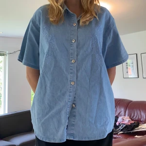 Ljusblå jeansskjorta med broderi  - Ljusblå jeansskjorta med kort ärm och avslappnad passform. Skjortan har broderade detaljer framtill med bladmönster och knäpps med silvriga knappar. Det står att skjortan är i storlek XXL och jag är vanligtvis storlek M ((jag TYCKER att den kanske snarare är en L, men det beror ju på hur man villa att den ska sitta:))