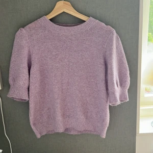 Ljuslila stickad tröja Vero Moda - Superfin ljuslila stickad tröja från Vero Moda med korta puffärmar och ribbade muddar. Perfekt för dig som vill ha en mjuk och mysig look med lite volym i ärmarna. Enkel rund halsringning och relaxed fit.