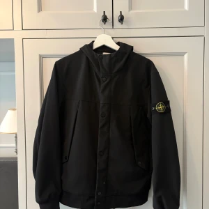 Svart regn/höstjacka från Stone Island - Svart softshelljacka från Stone Island med ikonisk patch på ärmen. Jackan har huva, knäppning med både knappar och dragkedja samt två snedställda fickor fram. Tillverkad i återvunnen polyester med vattenavvisande och andasfunktion. Perfekt för dig som gillar stilren och sportig look. Kostar ca 6400 ny
