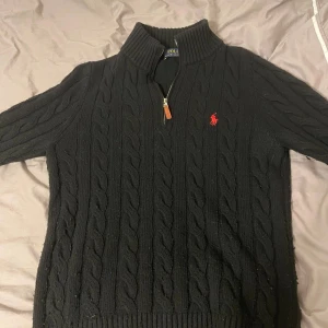 Svart stickad Ralph Lauren half-zip - Extremt bekväm och skön att ha på sig, den har en svart kabelstickad design som ser riktigt snyggt ut med den klassiska röd broderade loggan på bröstet och ett snyggt läderdetalj på dragkedjan. Ett perfekt val för dig som vill vara bekväm och stilig inför dem kyliga dagarna. 