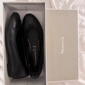 Svarta ballerinaskor från Tamaris - Stilrena svarta ballerinaskor från Tamaris i skinn med klassisk rund tå och söt rosettdetalj framtill. Perfekta för en enkel och snygg look, levereras i originalkartong. Använda endast en gång. Nypris 700kr. 
