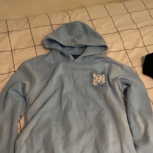 Ljusblå hoodie från Amiri - Säljer en ljusblå hoodie från Amiri med vit logga på bröstet och stor AMIRI-text på ryggen. Hoodien har huva, känguruficka och är tillverkad i mjuk bomull. Perfekt för dig som gillar streetwear och vill sticka ut med en snygg detalj på ryggen.