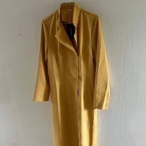 Senapsgul kappa  - Stilren långklänning i senapsgult från Woya med långa ärmar och knappar hela vägen framtill. Klänningen har en rak passform och hög krage, perfekt för dig som gillar minimalistisk och trendig stil.