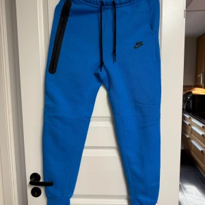 Blå Nike Tech Byxor - Snygga blå Nike Tech mjukisbyxor från Nike med svart snörning i midjan och en svart dragkedjeficka på sidan. Byxorna har smal passform och är gjorda i mjukt bomullsmaterial. Perfekta för chill eller träning, med klassisk Nike-logga på låret.