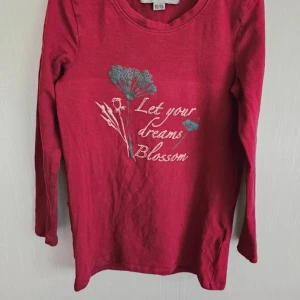 Vinröd långärmad tröja Amy & Lucy - Vinröd långärmad tröja från Amy & Lucy med tryck framtill som säger 'Let your dreams Blossom' och blommotiv i vitt och turkost. Tröjan har rund halsringning och är gjord i mjuk bomull, perfekt för en chill och snygg vardagslook. Sträcket är ifrån när den hängde på tork
