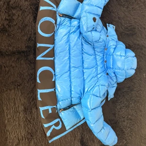 Blå Moncler pufferjacka - Snygg blå pufferjacka från Moncler med stor logga i ljusblått på svart panel nedtill. Jackan har huva, dragkedja, fickor med dragkedja och en ärmficka med Moncler-märke. Perfekt för kalla dagar och riktigt stil ren. En unik moncler jacka som är 100% äkta och köpt på NK förra året då jag fick den i present. Den är i storlek barn 164 men passar mig som brukar vanligt vis ha xs/s i kläder. Den är även unisex. Priset är inte hugget i sten, om ni har funderingarna är det bara att skriva 