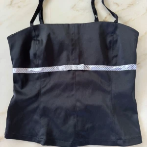 Svart korsettopp med prickigt band - Svart korsettopp med smala axelband och ett vitt band med små svarta prickar tvärs över bröstet. Bandet har en liten rosett i mitten som extra detalj. Snygg och stilren modell med figurnära passform.