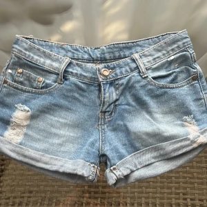 Ljusblå jeansshorts från JFR Brand - Snygga ljusblå low waist jeansshorts från JFR Brand med slitna detaljer framtill och nitar på ena bakfickan. Klassisk femficksmodell med uppvikta benslut och normal passform. Perfekta för sommardagar när du vill ha en chill och trendig look.
