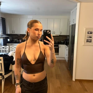 Brun faux skinn korsett-topp - Säljer en brun korsett-topp i faux skinn med smala halterneck-band och djup v-ringning. Toppen är croppad och har dragkedja i ryggen för enkel på- och avtagning. Storlek S