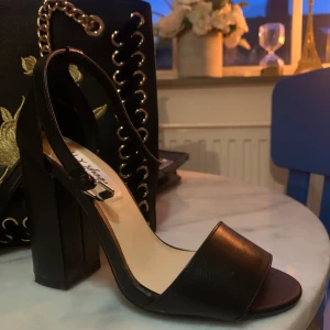 Svarta klackade sandaler med öppen tå - Stilrena svarta sandaler med hög klack och öppen tå. Skon har en bred rem över tårna och justerbart spänne vid ankeln. Perfekt för dig som vill ha en klassisk och trendig look. Tillverkad i skinnimitation med stabil blockklack.