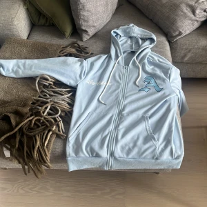 Ljusblå hoodie med Los Angeles-tryck - Säljer en ljusblå hoodie från ROMWE i storlek S. Tröjan har huva med snörning, dragkedja framtill och stora tryck med bokstaven A och texten Los Angeles på både fram- och baksidan. Tillverkad i 100% polyester och har långa ärmar.