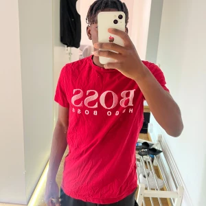 Röd Hugo Boss t-shirt med logga - Snygg röd t-shirt från Hugo Boss med stor vit BOSS-logga framtill. Klassisk passform med rund hals och korta ärmar. Tillverkad i mjuk bomull som känns skön mot huden. Perfekt för dig som gillar stilrena och trendiga plagg med tydlig branding.