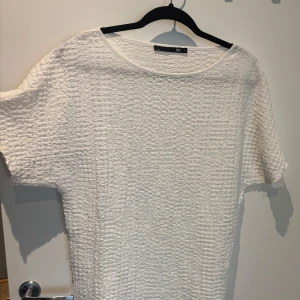 Vit strukturerad t-shirt  - Vit t-shirt från lager157 i modellen CLARA. T-shirten har en strukturerad, krinklad yta och är tillverkad i ett lätt material. Den har korta ärmar och en rund halsringning, perfekt för en clean och fräsch look.