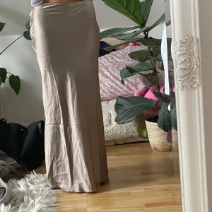 Beige långkjol i satin - Supersnygg beige långkjol med slät, glansig satin-feel. Kjolen har en figurnära passform upptill och faller rakt ner mot golvet. Perfekt för dig som gillar en clean och stilren vibe. Passar till både sneakers och klackar.