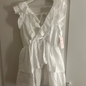 Vit volangklänning från Nelly - Supersöt vit kortklänning från Nelly med volanger, omlottliv och knytning i ryggen. Ärmlös modell. Aldrig använd lappen kvar! 