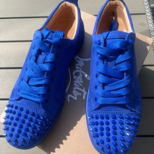 Blå Christian Louboutin sneakers med nitar - Snygga blå sneakers från Christian Louboutin i mocka med matchande blå snörning och coola nitar på tån. Skorna har den ikoniska röda sulan och en rund tå. Perfekta för dig som vill sticka ut med en lyxig och edgy stil, orginal förpackning tillkommer 