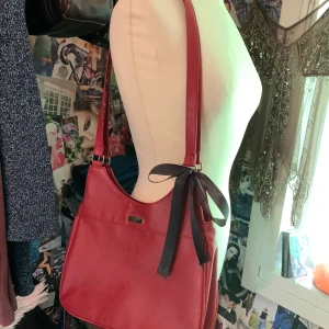 Röd axelväska från Eternal Line - Snygg röd axelväska i skinnimitation från Eternal Line. Väskan har en svart rosett på axelremmen som man kan juster/ta bort och en praktisk dragkedja på baksidan. Perfekt storlek för att få plats med allt du behöver och stilren design som sticker ut.