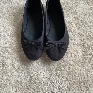 Svarta ballerinaskor med rosett - Stilrena svarta ballerinaskor med diskret struktur i tyget och en söt rosett framtill. Skorna har rund tå och platt sula, perfekta för en enkel och klassisk look. Tillverkade i textilmaterial som känns lätta på foten.