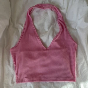 Rosa ribbad croppad linnetopp - Säljer en rosa croppad linnetopp med ribbad struktur och djup v-ringning. Toppen är ärmlös och har halterneck-modell som knyts i nacken. Perfekt för varma dagar och enkel att styla med jeans eller kjol