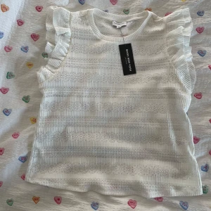 Vit blus med volang från Marc New York - Supersöt vit blus från Marc New York med genombrutet randigt mönster och volangdetaljer på axlarna. 