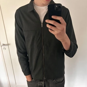 Svart overshirt med dragkedja - Stilren svart overshirt med rak passform och dragkedja framtill. Jackan har klassisk krage, en bröstficka och långa ärmar. Perfekt att slänga över en t-shirt för en clean look. Storlek 48 så som en M