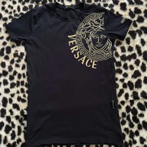 Svart - Designer T-Shirt - Svart t-shirt med stort guldigt Medusa-tryck och logga på bröstet. Klassisk rund hals och korta ärmar. Snygg och stilren design som sticker ut med den ikoniska looken. Tröjan är i nyskick, och priset går att diskutera vid snabb affär🏆 Kom i DM om ni vill ha fler detaljer om märket😇